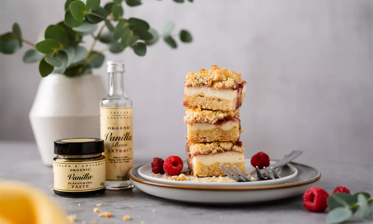 Šviesiame keraminiame dubenyje sluoksniuotas trapus pyragas su trupiniais, avietėmis šalia, dekoratyvines šakelės ir vanilės ekstrakto buteliukai.