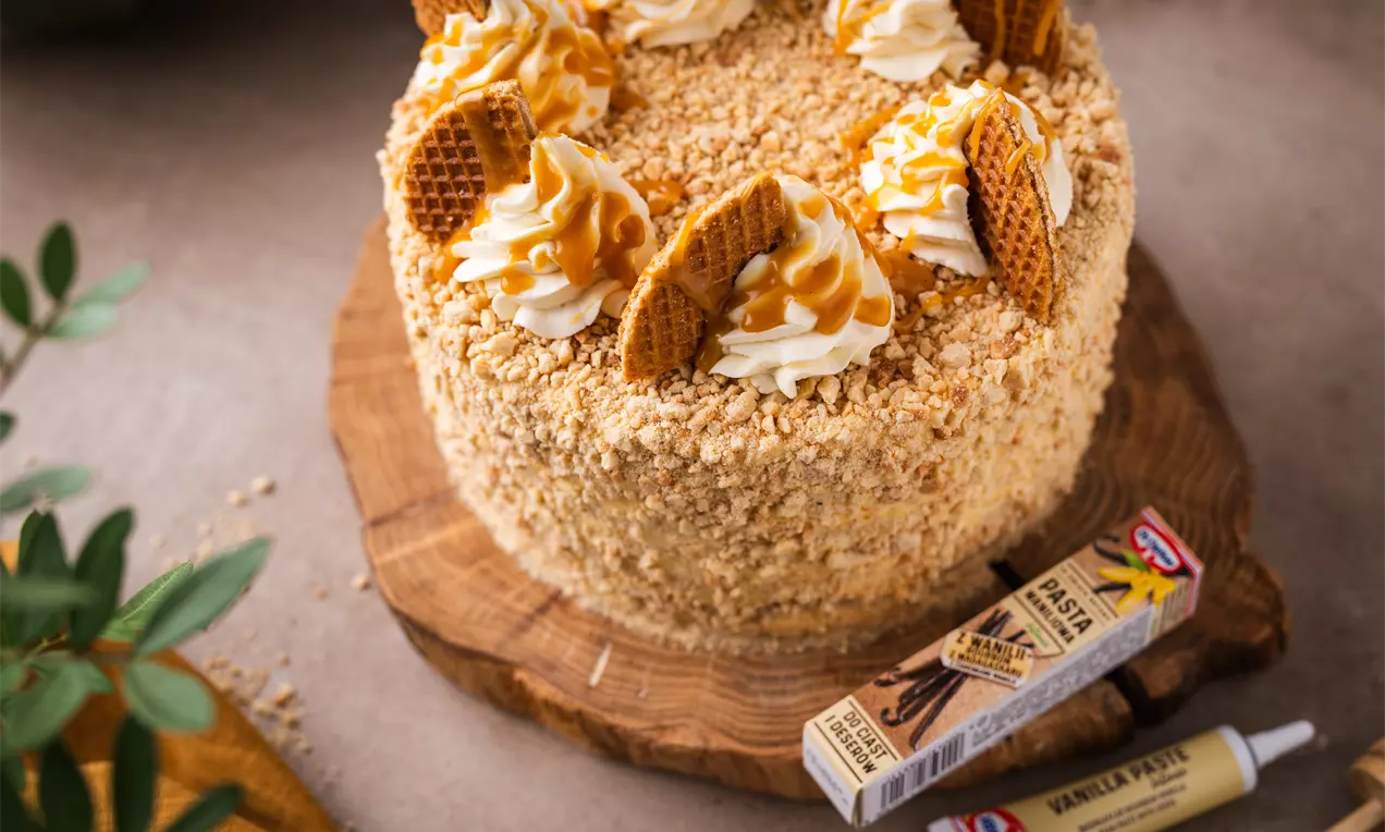 Auksinės karamelės spalvos purus tortas, apibarstytas trupiniais, puoštas grietinėlės kepurėlėmis ir vafliniais gabalėliais ant medinės lėkštės