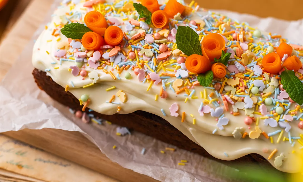 Pailgas morkų keksas su šviesiai kreminiu glajumi, dekoruotas spalvotomis pabarstomis, morkų suktinukais ir šviežiais mėtų lapeliais