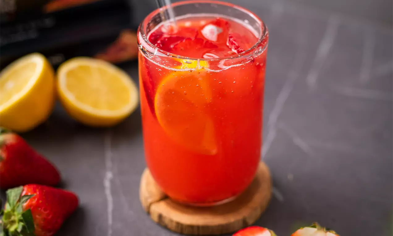 Skaidrus stiklinis indas su ryškiai raudonu limonadu, dekoruotas citrinos griežinėliu, plonu šiaudeliu ir šviežiomis braškėmis šalia